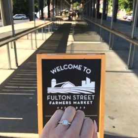 Close-up of an engagement ring in front of the Fulton Street Market welcome sign in Grand Rapids, MI